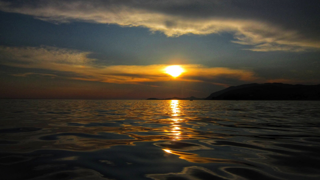 This image captures a breathtaking sunset over a calm body of water. The sun is partially obscured by scattered clouds, casting a warm, golden glow across the horizon. The reflection of the sunlight shimmers on the gently rippling water, creating a serene and tranquil atmosphere. In the distance, silhouettes of hills or mountains are visible, adding depth to the scene. The sky is a blend of soft oranges, yellows, and deep blues, enhancing the peaceful ambiance of the setting sun. The overall mood of the image is calm, reflective, and picturesque.