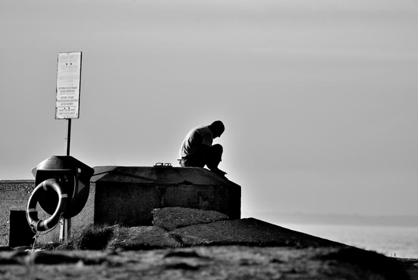 Une photo en noir et blanc au format paysage. Ciel clair et ligne d'horizon au quart inférieur de l'image. Sur ce fond, se détache la silhouette massive d'un bunker en partie enterré, on voit sur la gauche une entrée tournant le dos à la mer. Un panneau  documente le lieu et une bouée semble rattachée à un contenant métallique. Au centre de l'image, un homme vu de profil est assis sur le toit en béton, genoux replié et tête baissée, seul.
