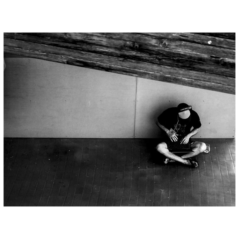 This black-and-white image shows a person sitting alone on the ground, leaning against a wall under a wooden structure. The individual is using a laptop, which is placed on their lap. The person is wearing a cap, a dark t-shirt, headphones and shorts. The scene is captured from an elevated angle, emphasizing the solitude and focus of the person in a quiet setting. (draft by mistral.ai, revised by author)