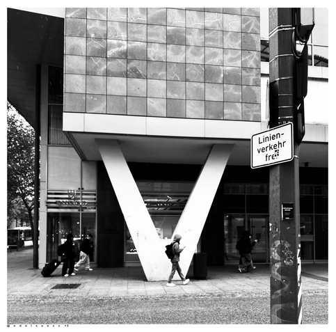 Schwarzweißfotografie: Innenstadt, zubetoniert, Fußweg vor Hausfassaden, eine helle V-förmige Abstützung aus Beton trägt ein kastenförmigen Überbau aus den 1970er Jahren, Passanten laufen auf dem Fußweg, an einem Mast steht ein Schild mit der Aufschrift: „Linienverkehr frei“  