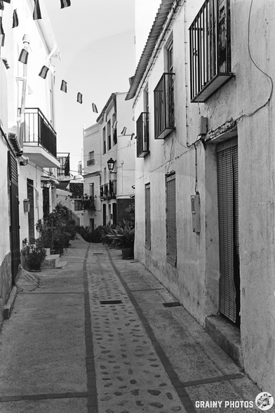 A narrow, empty street in Guájar-Faragüit, a Mediterranean village lined with white buildings, balconies, and potted plants. Flags hang across the street and a lantern is mounted on a wall. The photo is black and white.