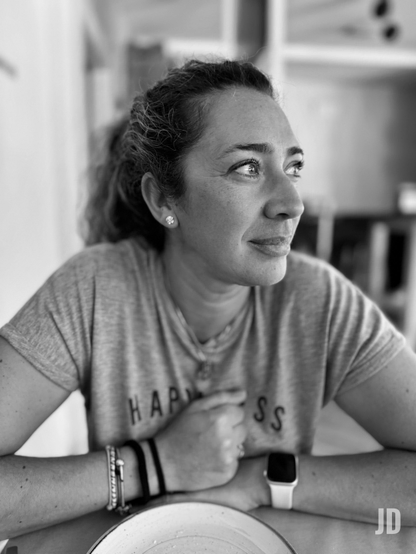 Una fotografía en blanco y negro de una mujer sentada en una mesa, mirando pensativamente hacia la derecha con una leve sonrisa. Tiene el cabello oscuro recogido y lleva pendientes pequeños de botón. Viste una camiseta gris de manga corta con las palabras "HAPPINESS" parcialmente visibles en el pecho. En su muñeca izquierda, lleva varias pulseras, y en la derecha, un reloj inteligente de banda blanca. Sus brazos están apoyados en la mesa, donde se ve parte de un plato en primer plano. El fondo está desenfocado, sugiriendo un interior. La imagen tiene un enfoque nítido en su rostro y expresión tranquila.