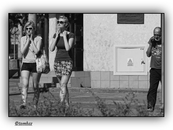 Black-and-white street photograph in full sunlight. On the left side of the frame, two young women walk side by side. The first, wearing a light blouse and dark shorts, with her hair tied back and sunglasses on, touches her neck with both hands — a delicate, almost instinctive gesture. The second, in a dark top and patterned skirt, also wearing sunglasses, mirrors the gesture — hands near her neck, the movement similar but not identical. Their posture is upright, their stride confident, their physical presence clear. On the right side of the frame, an older man in dark clothing walks alone, a plastic bag slung over his shoulder. His face is tense, gaze distant, posture slightly hunched — weariness visible in every gesture. The background features a tiled wall, a commemorative plaque, and an electrical box with a warning symbol. In the foreground, blurred blades of grass. The contrast between youth and age plays out in the rhythm of bodies and gestures.