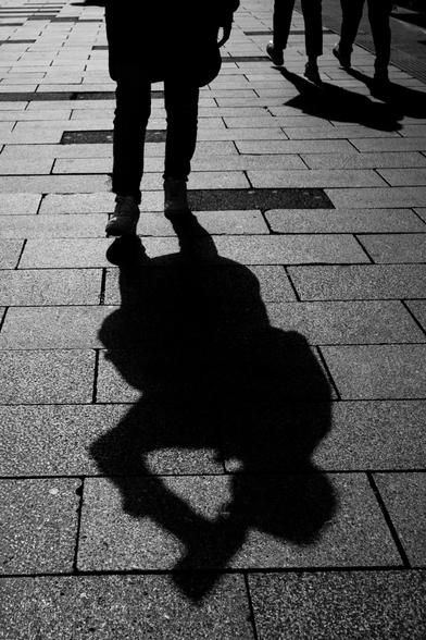 The image is a black and white photograph taken from a low angle, focusing on the legs and shadow of a person on a stone-paved surface. 

The main subject is the shadow cast by the person, which is a large, dark silhouette dominating the lower half of the frame. The arms are positioned as if the person is drinking something. The shadow's form is sharply defined, indicating a strong light source from above.

Above the shadow, the person's legs are visible from the knees down. 