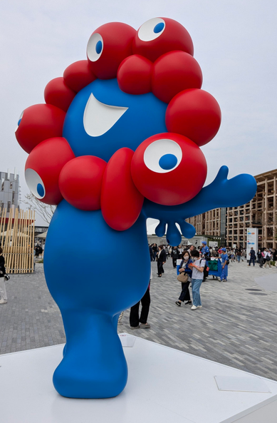 Eine blaue Figur. Sie lacht und ein Arm ist zur Seite ausgestreckt.. Der Kopf ist mit roten Kugeln umgeben, welche wohl Augen darstellen sollen. 