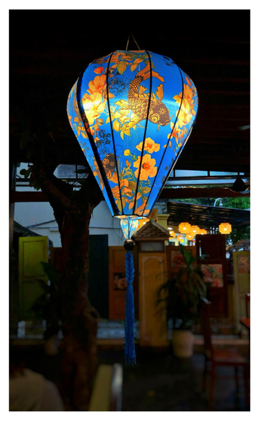 Photo of an illuminated Vietnamese lantern suspended from a dark ceiling in a hotel’s covered lobby. The lantern is shaped like a hot air balloon, with silk fabric stretched over bamboo ribs. It features a design of stylized orange-and-black koi fish and orange flowers on a vivid blue background. A long blue tassel hangs from the lantern bottom. Out of focus beyond the lantern are a slim tree trunk, tables and chairs, and a barrier made of doors of different sizes and colors joined together. Outside is a glimpse of orange lanterns hanging in the covered lobby of another hotel.