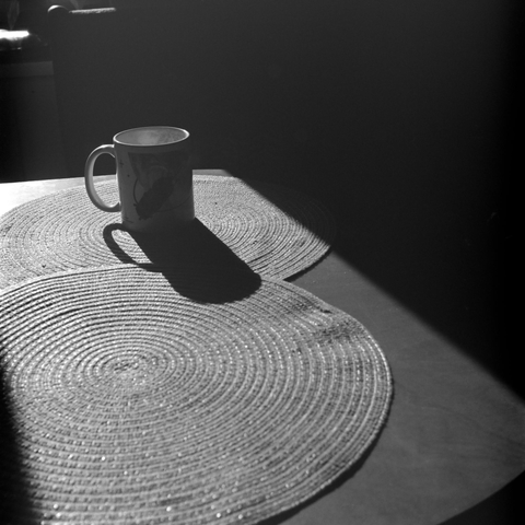 The photograph shows a mug on a wicker stand. The mug has a handle, which casts a shadow. The photograph is black and white, giving it a minimalist yet artistic look.