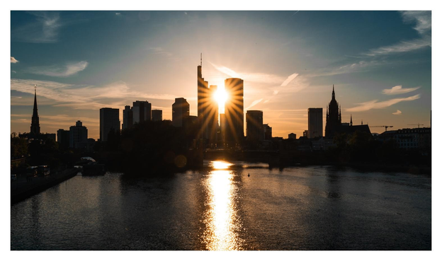 Skyline von Frankfurt als Schwarze Silhouette vor abendlichem Himmel. Zwischen den Hochhäusern strahlt ein Sonnenstern hindurch.