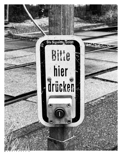Schwarzweißfotografie: Drücker an einem Straßenbahnübergang, Hinweis ausgeschrieben auf einem Schild, an einem Mast: „Bitte hier drücken“ darunter ein Taster, dahinter unscharf Straßenbahnschienen, Betonplatten dazwischen 