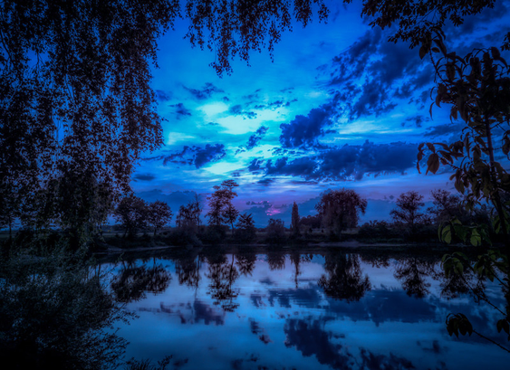 Ein Blick zur Blauen Stunde über einen kleinen See. Dieser ist mit Bäumen am Ufer gesäumt die, wie auch die Wolken und der Himmel, sich auf der Wasseroberfläche spiegelm. Im Vordergrund sind die Äste von zwei Bäumen zu sehen die das Bild einrahmen. Am Horizont ist noch ein kleiner Flecken der violett leuchtet zu sehen, der von der untergegangenen Sonne stammt.