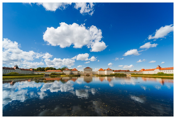 Ultraweitwinkelaufnahme eines breiten Schlosskomplexes vor einem quasi spiegelglatten See. Im See spiegeln sich Schloss und der blaue Himmel.