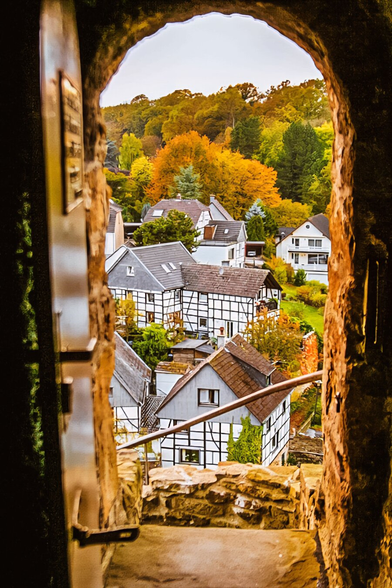 A picturesque view through a stone archway reveals a charming village with traditional half-timbered houses surrounded by vibrant autumn trees in shades of gold, orange, and green.