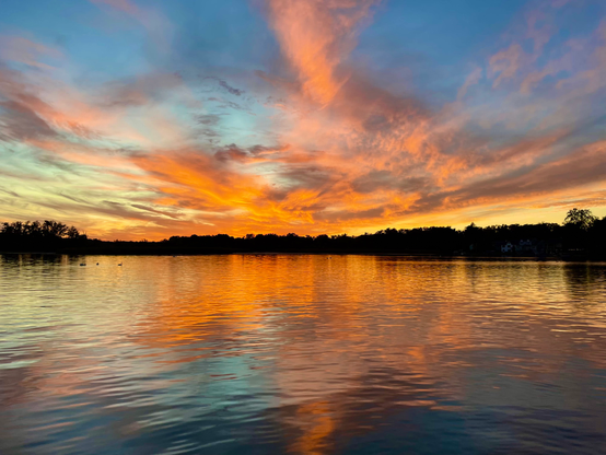 Sunset photo of bright orange and peach clouds reaching out from the horizon and reflected in a lake.