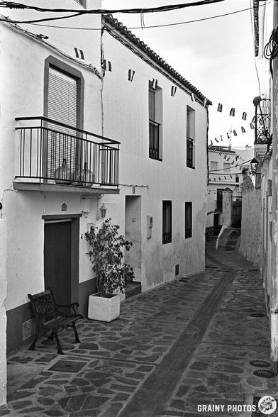 Black and white photo of an empty narrow cobbled street in Guájar-Faragüit, lined with white buildings, a balcony, a bench, potted plants, and bunting overhead.