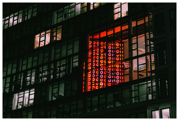 Blick auf eine Glasfront eines Bürogebäudes von schräg unten. Hinter einem der großen Fenster steht eine Lichtinstallation aus vielen in einem Raster angeordneten rot glühenden Doughnut-Förmigen Lampen.
