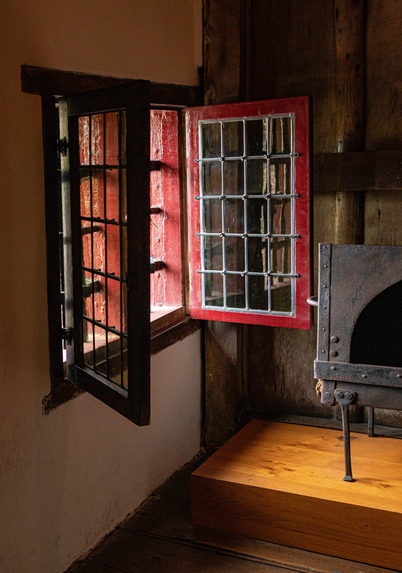 Photo of a corner of a room, with a white plastered wall on the left containing an open window. The window is split into two sections that open inward. These two windows have red-painted wooden frames and stained glass panes. The windows themselves have crosswise iron round mullions. The light shines into the room. On the right, part of an iron wood-burning stove is visible.