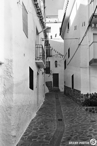 Narrow cobbled street in Guájar-Faragüit, lined with whitewashed buildings and balconies, captured in black and white. Flags hang above the alleyway, and a closed door is visible at the end of the street.
