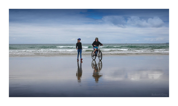 Two children on a beach, one riding a bicycle, walk towards the camera with the sea behind them. The weather is overcast and the colours are muted but with high contrast.