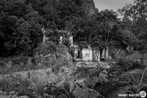 Black and white photo of a crumbling, abandoned mill near Guájar-Faragüit, Spain, overgrown with vegetation and surrounded by trees and rocky terrain. The weathered structure has  partially collapsed.