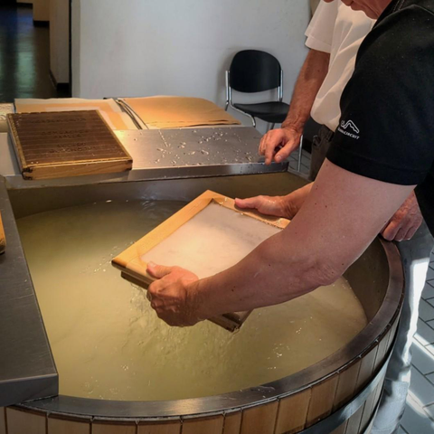 Gezeigt wird das Papier schöpfen.
Zwei Arme heben einen Holzrahmen, bespannt mit einem feinen Netz, aus einem großen Trog mit Wasser und Zellulose.