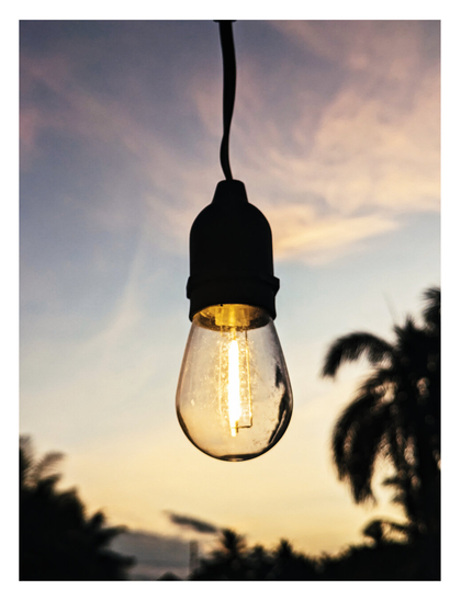 A lit outdoor light bulb hanging from a dark wire against a sunset sky.