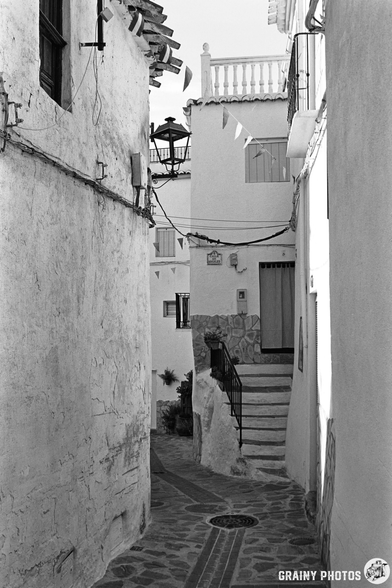 Black and white photo of a narrow, winding street in Guájar-Faragüit, nestled between old white buildings with textured walls and stone steps, decorated with small hanging flags. Sunlight casts shadows across the quaint, quiet alleyway.