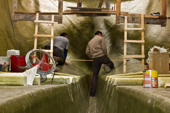 The photo is taken from the inside of a boat under construction, towards the bow. The inside of the boat is covered with a light yellow cloth, stretched tightly along the wall. To the left and right are raised platforms, and in the center is a walkway that narrows towards the front due to the perspective. Two men sit in the bow (center of the photo). They hold a long measuring stick. To the left and right of the men are two wooden ladders leaning against a wooden structure attached somewhere at the top. Various pots and other materials are placed on the raised platforms to the left and right.