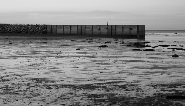 Black and white photograph of a beach at low tide with a stone and wooden dock jutting out into the water, with a person walking on it wearing dark trousers and a white sweater, looking at their phone. The sea is visible to the right at the end of the dock. At the front of the dock, the rippled sand is still covered in some water in places, reflecting the light. Seagulls, stones, and piles of seaweed are scattered here and there.