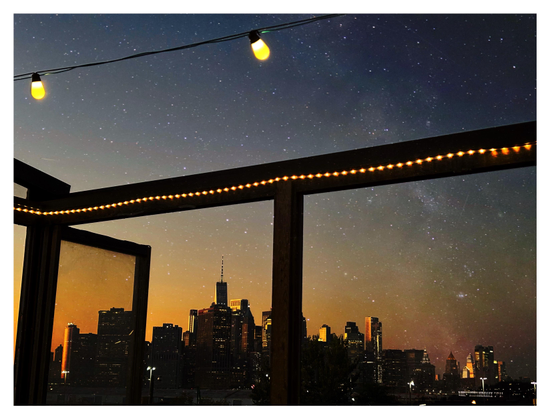 Wishing my city had less light pollution, I made this photo montage of an impossible view: a sky dense with stars over the lower Manhattan skyline against the orange glow of sunset, as seen from a rooftop restaurant in Brooklyn. (The sunset and city lights would normally wash out the stars.) The skyline — building silhouettes dotted with lit windows — is seen through the frame of the restaurant’s roofless wooden structure, with a string of lights lining the upper horizontal beam. A couple of larger lightbulbs are suspended from wires stretching across the sky overhead. The starry night sky (with a hint of the Milky Way) was photographed in northern Idaho.
