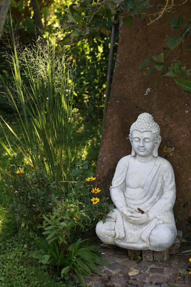 Eine weisse Statue, eines sitzenden Buddhas vor einem Sandsteinobelisk. Links von der Statue blühen drei gelbe Mageriten. Weiter links ragen Gräser empor und werden leicht von der Sonne durchstrahlt. Von oben rechts ragen ein paar Blätter Efeu in das Bild hinein.