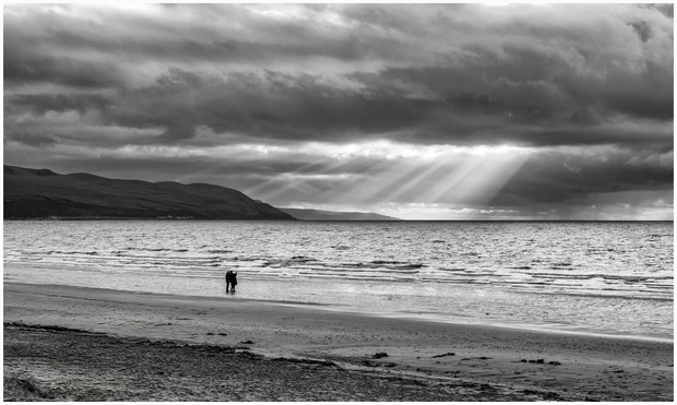 A couple walks hand in hand along a beach, with gentle waves lapping at their feet. The scene is set against a dramatic sky filled with dark clouds and rays of light streaming down. The image is in black and white, enhancing the atmospheric feel.