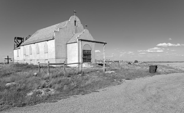 A black and white landscape photo of an abandoned church with a graveyard just beyond. The church is on the left side of the frame and it faces toward the right. The front door is boarded up as are the five windows on the right side of the building. There's a short in height entrance with an cross on top and then a taller wall behind the front entrance area that has the bell tower and a cross on top. The building is Spanish mission style. The building is a light color and the roof is dark. There's a tall cross on the ground immediately behind the church. In front there's a tall wood entrance where a gate likely once was and there are three short sections of fence on each side of the gate framework. A large commercial-sized rusted metal trashcan sits on the right next to the gravel drive in the foreground. The graveyard is seen on the slight hill behind and to the right of the church building. There's a few clouds low on the horizon otherwise the sky is clear.