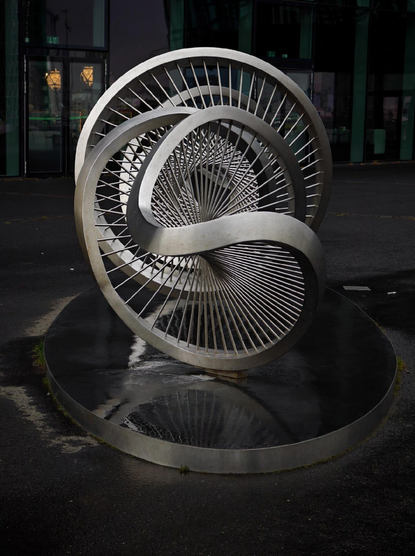 A photo of a metallic abstract sculpture with interwoven curved elements connected by smaller elements like spokes. It stands on an oval reflective base outdoors in front of a dark building in the background.

