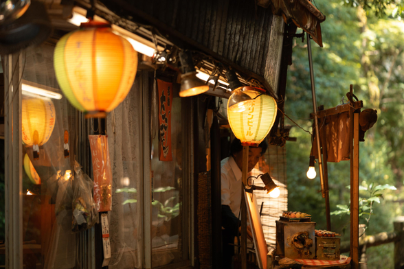 A small shop at the side of a hiking trail leading to Minoh Falls, close to Osaka in Japan. The photo shows the shop windows with glowing lanterns and a small grill in front of greenery