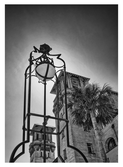 Black and white photo of a vintage streetlamp in the foreground, framed by tall historic stone towers and a palm tree under a clear sky. Majestic and timeless.