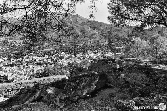 Black and white photo showing a rocky foreground with trees, overlooking a hillside town with many buildings and mountains in the background.