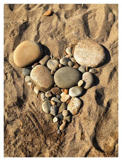 Photo looking down at a collection of assorted, differently sized smooth rocks on a sandy beach, arranged in the shape of a heart. Sunlight brings out the highlights, shadows, colors and textures of the rocks, as well as of the scoops and hollows in the sand.