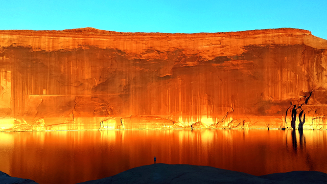 A sandstone wall on the other side of calm lake water, the wall is bathed in sun, shadow in front, a person is standing in the shadow.
