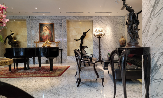 White marble floors and walls, a black grand piano to the left, the painting behind the piano is of Jesus holding a lamb. center are low seat chairs, at the fall wall is a black statue of what looks like a lady running. To the right on a tall black table is a French bronze woman in robes holding a lamp a cupid is at her feet 