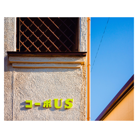 A color photo of a beige stucco building with a latticed window above and bright green letters on the wall reading “コーポUS.” The sunlight casts warm shadows across the textured surface, and a clean blue sky fills the right side of the frame.