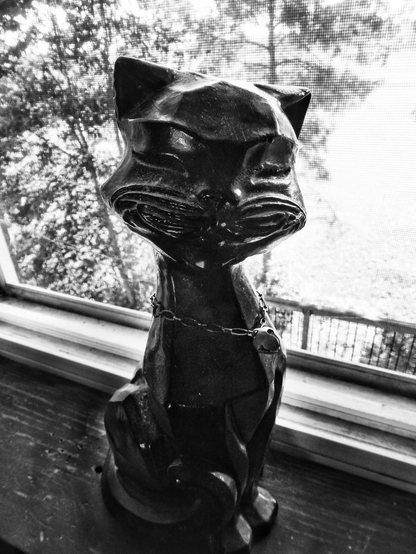 A black ceramic cat sits in front of a window. The cat's face is pointed towards the camera and its whiskers and eyes can be seen. A large tree is shown through the window in this black and white image.