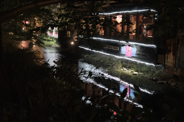 Photograph of a water stream bordering the back wall of a restaurant at nighttime, with the neon lights surrounding the restaurants' windows reflected in the stream water. Through the windows, the warmly-lit interior of the restaurant can be seen, including patrons and a waitress. The view is seen through the dark yet sparce foliage of trees besides the stream.