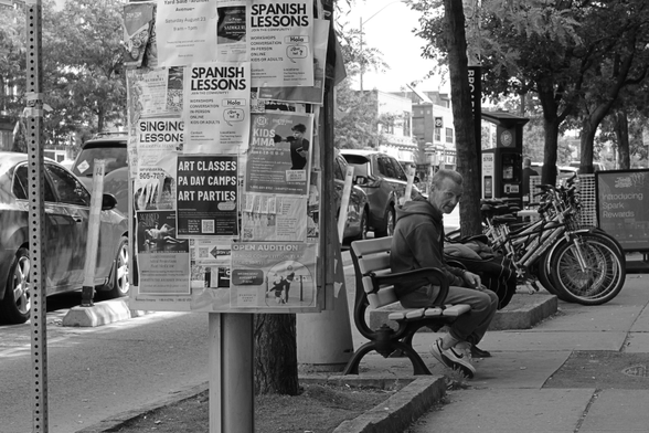 Just left of centre, a neighbourhood poster board fills the frame, covered in printed posters offering language lessons and art classes. Beyond it, an older man sits on a bench, looking at the camera. Further in the distance, toward the right of the frame, are several bicycles parked in a neat row.
