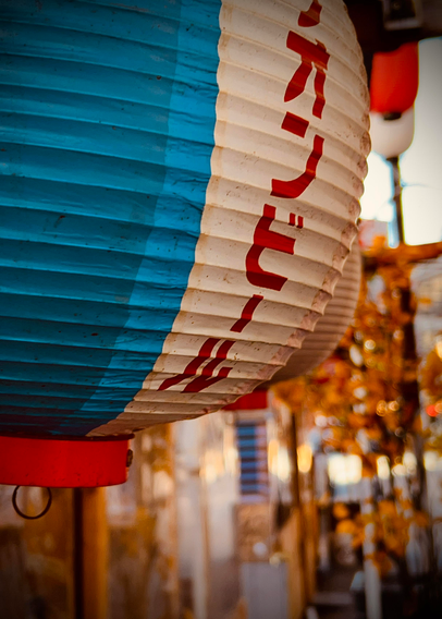 A color photograph in portrait orientation. A close-up, slightly low-angle photograph of a large, cylindrical Japanese paper lantern. The lantern is primarily white and blue. The left half is solid teal-blue, and the right half is white with large, bold, red Japanese characters written vertically down the center in Katakana script. The lantern has a bright red base and. A small black metal ring is visible attached to the red base. The paper texture of the lantern is ribbed. The background is softly blurred, featuring warm, golden-yellow bokeh lights and indistinct shapes of the market in the background. Another similar lantern, slightly out of focus, is visible behind the main one.