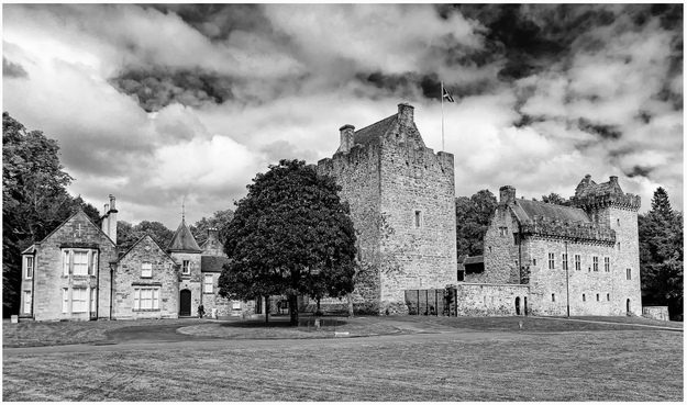 A historical stone castle complex surrounded by greenery and a lawn, captured in black and white. The scene includes multiple buildings with distinct architectural features and a cloudy sky above.