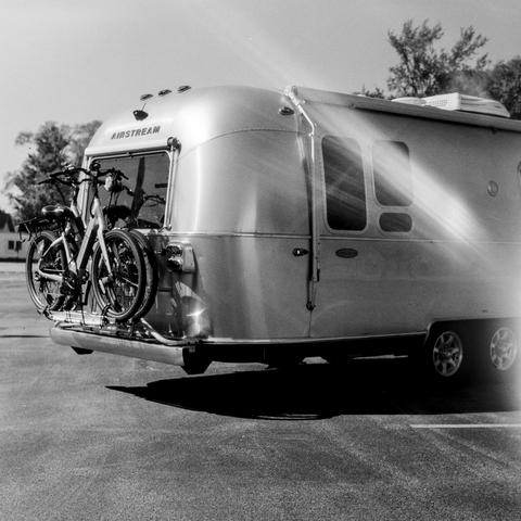 Black and white photo of a classic silverr Airstream camper is show from the back right and only about 70% of the length of the campter is in frame. The camper is in a parking lot under a bright sun and the sunlight is gleaming off the smooth curves of the sides and top. The back of the camper hsa two step-through electric bicycles. The whole vibe is ready for summer adventures. There's a light leak in the photo that looks like a stylistic flare across the right side of the camper.