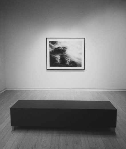 A black and white image from inside an art gallery. A large picture hangs on a white wall, and a large rectangular bench sits on a wooden floor in front of it.