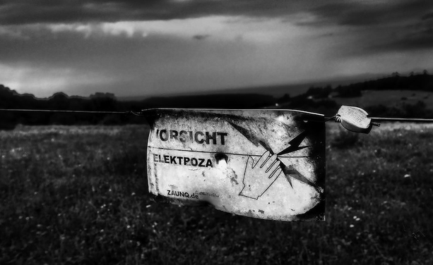 E9n elektrozaun für Rinder. Ein Schild hängt daran was ziemlichdeformiert ist und nur unleserlich auf diesen Stromzaum hinweist. Im Hintergrund kann man unscharf die Wiese und einige Bäume erkennen. Das Foto ist in Schwarzweiß.