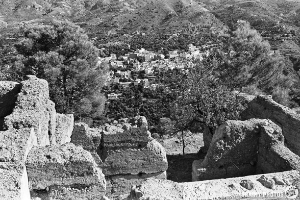 Black and white photo showing the abandoned Castillejo near Guájar-Faragüit, its ancient stone ruins in the foreground, with trees and a distant village nestled among hills and greenery in the background.