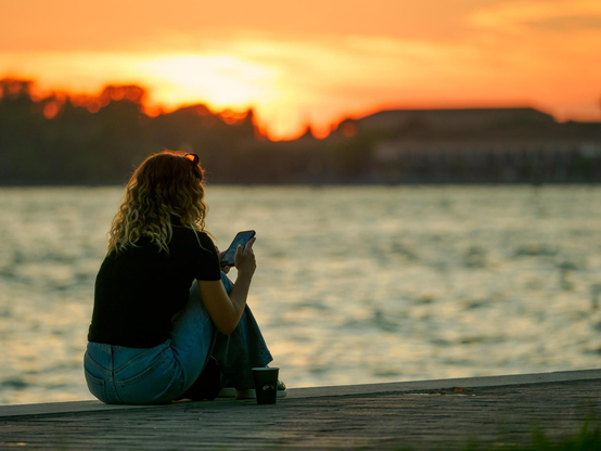 A woman with curly hair sits cross-legged on a wooden pier at sunset, looking at her smartphone, with a coffee cup beside her and a calm body of water in the background.